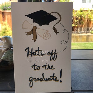May include: A graduation greeting card with a black mortarboard, diploma, and the text "Hats off to the graduate!" in navy blue script. The card has braille text above the script.