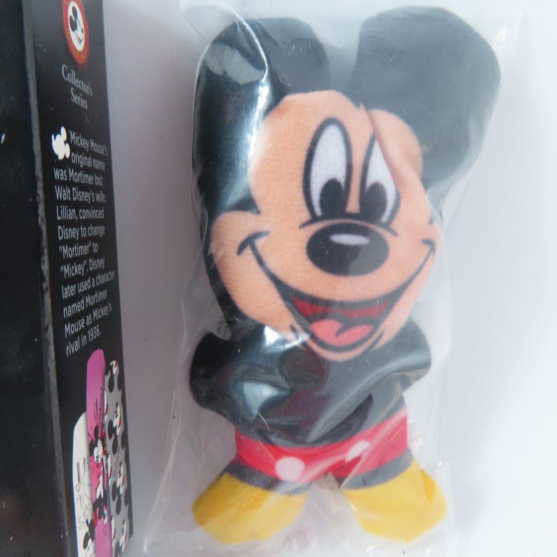 May include: A plush Mickey Mouse figure in a clear plastic bag, featuring black ears, a tan face, red shorts with white polka dots, and yellow shoes. Beside it is a black box labeled Collector's Series with text about Mickey Mouse's history.