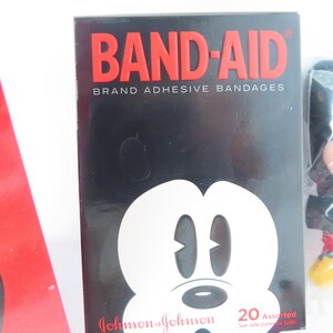 May include: A rectangular black box of Band-Aid brand adhesive bandages featuring a white graphic of a cartoon mouse face. The text reads BAND-AID BRAND ADHESIVE BANDAGES, Johnson & Johnson, and 20 Assorted. A red object is partially visible on the left.