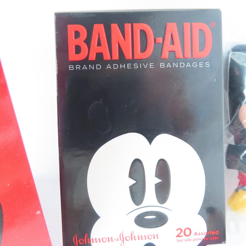 May include: A rectangular black box of Band-Aid brand adhesive bandages featuring a white graphic of a cartoon mouse face. The text reads BAND-AID BRAND ADHESIVE BANDAGES, Johnson & Johnson, and 20 Assorted. A red object is partially visible on the left.