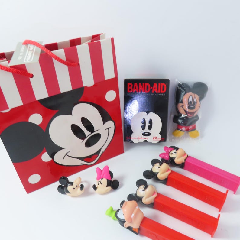 May include: A red and white striped gift bag featuring Mickey Mouse, a box of Band-Aid bandages, a small Mickey Mouse figure in plastic, two small character heads, and four Pez dispensers with Mickey, Minnie, and Goofy character heads.