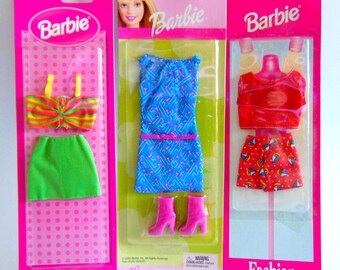May include: Three Barbie Fashion Favorites doll clothing sets in original packaging. The left set includes a striped crop top and green skirt. The centre set features a blue patterned dress and pink boots. The right set contains a red top and patterned shorts.