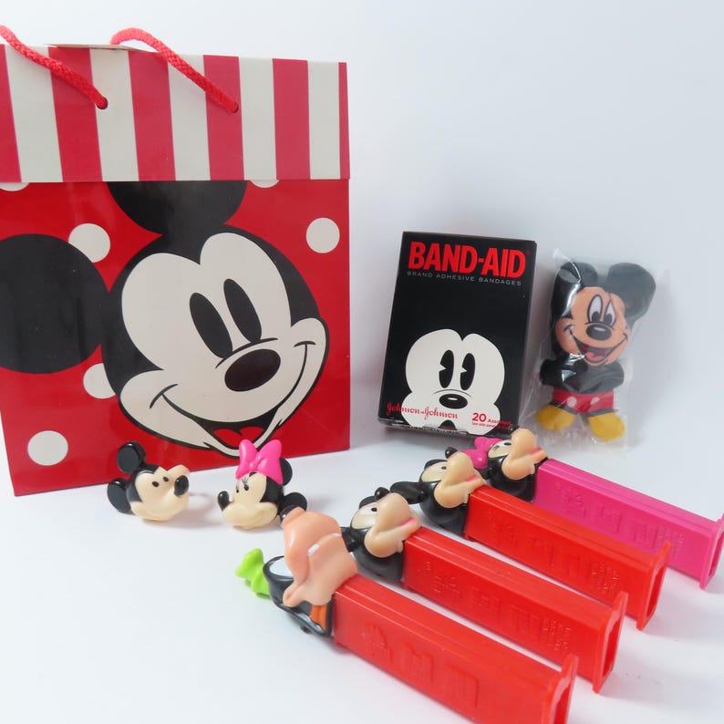 May include: A red and white striped gift bag with a Mickey Mouse graphic, a box of Band-Aid brand adhesive bandages, a small Mickey Mouse plush, two character rings, and three Pez candy dispensers featuring Mickey and Minnie Mouse figures.