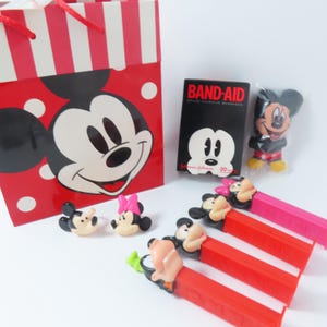 May include: A red and white striped gift bag featuring Mickey Mouse, a black box of Band-Aid brand adhesive bandages, a small Mickey Mouse figure in plastic, two small character heads, and four colorful Pez candy dispensers with character toppers.