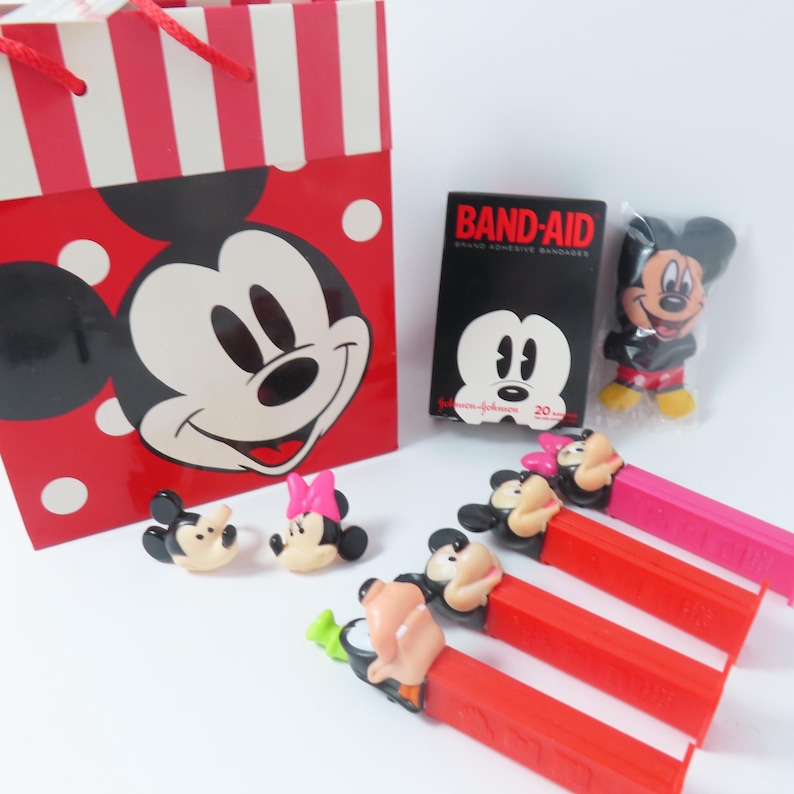 May include: A red and white striped gift bag featuring Mickey Mouse, a black box of Band-Aid brand adhesive bandages, a small Mickey Mouse figure in plastic, two small character heads, and four colorful Pez candy dispensers with character toppers.