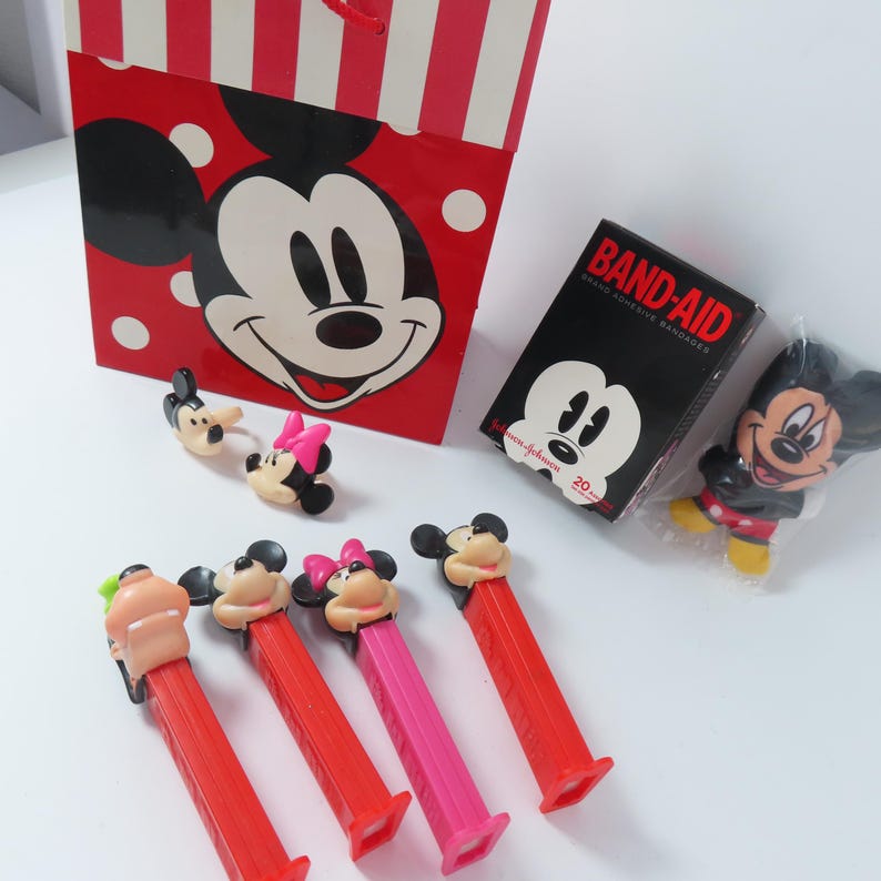 May include: A red and white striped gift bag featuring Mickey Mouse, a box of Band-Aid adhesive bandages, a small Mickey Mouse figure in plastic, two small character rings, and four colorful Pez candy dispensers with character heads.