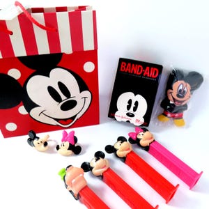 May include: A red and white striped gift bag featuring a Mickey Mouse graphic, a box of Band-Aid adhesive bandages, a plastic-wrapped Mickey Mouse figure, two small character rings, and four colorful Pez candy dispensers with character heads.