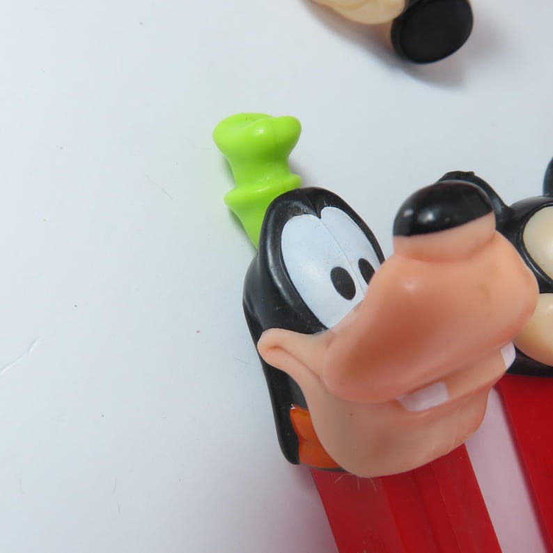 May include: A plastic Pez candy dispenser featuring the character Goofy with a black head, white eyes, a peach-colored snout, and a bright green hat. The dispenser body is red and rectangular, extending downward from the character head.