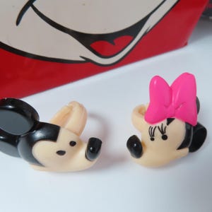 May include: Two plastic rings featuring character heads. One ring is a black and beige mouse head with round ears. The other ring is a beige mouse head with a bright pink bow and black eyelashes, positioned in front of a red and white graphic background.