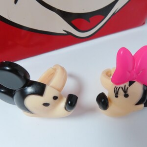 May include: Two plastic rings featuring the heads of cartoon characters. One ring depicts a black and beige mouse with round ears, and the other depicts a mouse with a bright pink bow, black eyelashes, and a beige face. Both have adjustable bands.