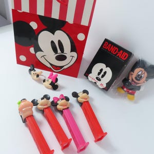 May include: A red and white striped gift bag featuring Mickey Mouse, a box of Band-Aid adhesive bandages, a small Mickey Mouse figure in plastic, two small character rings, and four colorful Pez candy dispensers with character heads.