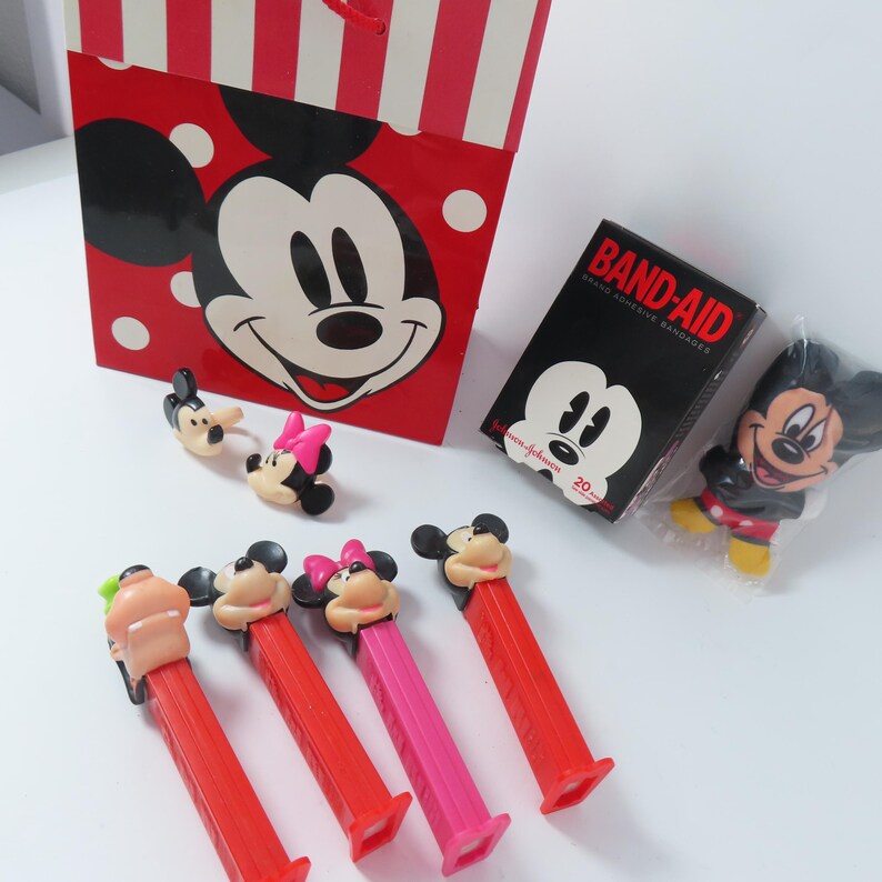 May include: A red and white striped gift bag featuring Mickey Mouse, a box of Band-Aid adhesive bandages, a small Mickey Mouse figure in plastic, two small character rings, and four colorful Pez candy dispensers with character heads.