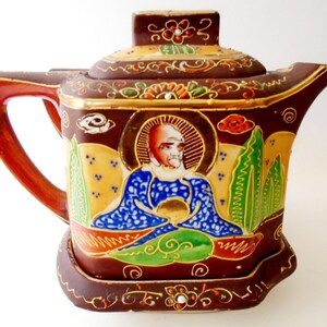 May include: A square-shaped ceramic teapot with a dark brown base color, featuring a hand-painted figure in a blue patterned robe surrounded by green trees and gold accents. The teapot includes a matching lid and a handle on the left side.