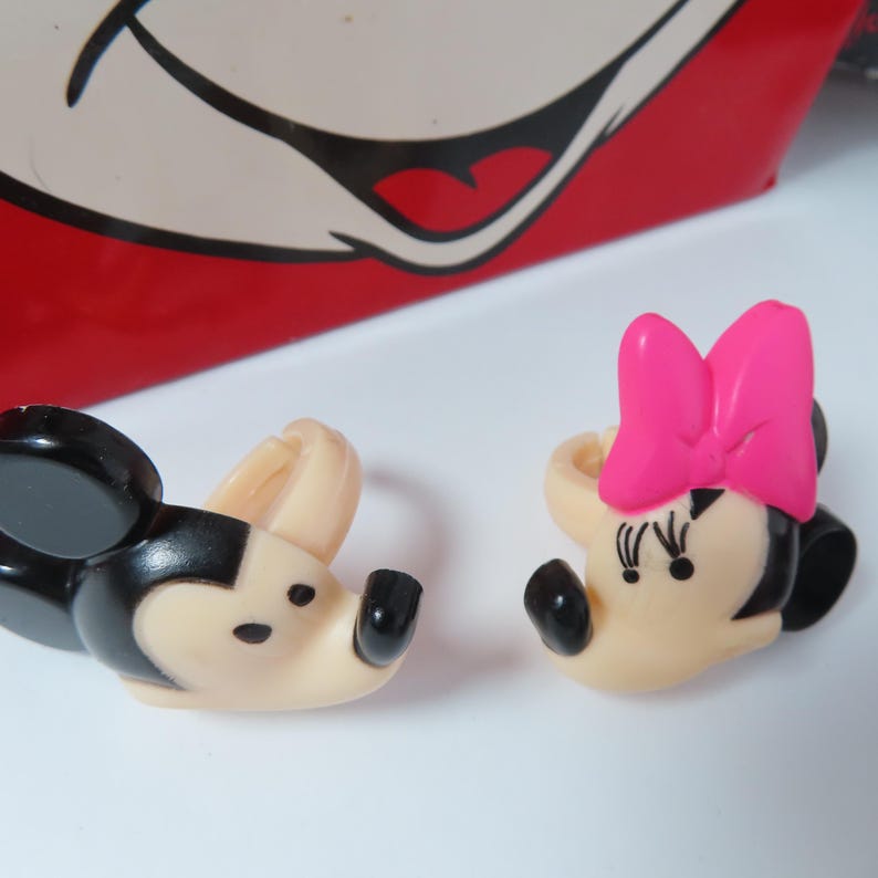 May include: Two plastic rings featuring character heads. One ring is a black and beige mouse head with round ears. The other ring is a beige mouse head with a bright pink bow and black eyelashes, positioned in front of a red and white graphic background.