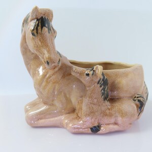 May include: A glossy ceramic planter shaped like a mare and foal lying down. The sculpture features a light tan glaze with dark brown painted details on the manes and hooves. The hollow center of the planter is positioned behind the horses.