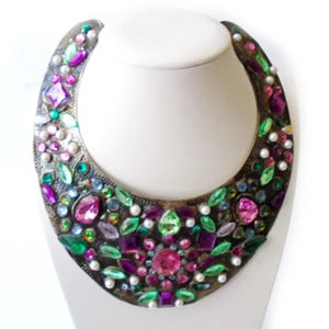 May include: A wide, crescent-shaped bib necklace featuring a dark, oxidized metal base adorned with an intricate arrangement of faceted pink, purple, and green crystals, along with small, round white faux pearls embedded throughout the surface.