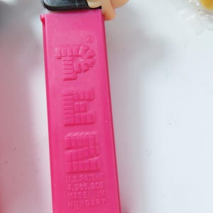 May include: A bright pink plastic Pez candy dispenser featuring a molded vertical logo on the front. The base includes the text U.S. PATENT 4.966.305 and MADE IN HUNGARY. A black plastic component is visible at the top of the dispenser body.