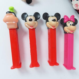 May include: Four plastic Pez candy dispensers featuring character heads. From left to right: Goofy with a green hat, two Mickey Mouse figures with black ears, and Minnie Mouse with a pink bow. Three dispensers have red stems, and one has a pink stem.