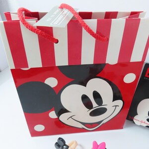 May include: A red and white gift bag featuring a large Mickey Mouse face graphic, white polka dots, and red striped top border. The bag has red rope handles and a rectangular tag attached that reads Stephen Lawrence, Cranbury, NJ, USA.