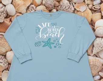 May include: A light blue long-sleeve shirt with the text "Life is Better at the Beach" in white script. The design includes a teal starfish and seashell graphic. The shirt is displayed on a background of seashells.