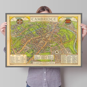 May include: A vintage map of Cambridge, England, with a person holding it up. The map is in a dark gray frame and features a detailed illustration of the city's streets, buildings, and landmarks. The map is titled "Cambridge" and includes a key for identifying colleges and churches.