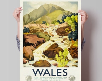 May include: A vintage travel poster advertising Wales with a scenic illustration of a waterfall cascading through a rocky landscape with green trees and a mountain in the background. The poster features the text "WALES" in bold blue letters and the logo of the Great Western Railway (GWR) in the bottom corners.