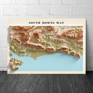Puede incluir: Un mapa topográfico enmarcado de South Downs Way, con una representación 3D detallada del paisaje. El mapa muestra tonos de marrón, verde y azul, con el texto "SOUTH DOWNS WAY" en la parte superior.