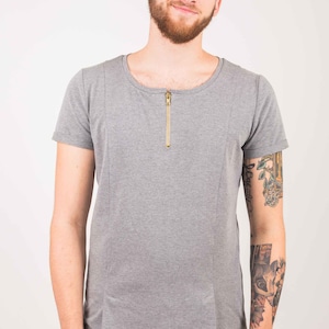 Könnte beinhalten: Ein graues T-Shirt mit kurzen Ärmeln und einem goldenen Reißverschluss am Ausschnitt. Das Shirt hat eine lockere Passform und eine etwas längere Länge. Das Model trägt eine schwarze Hose.