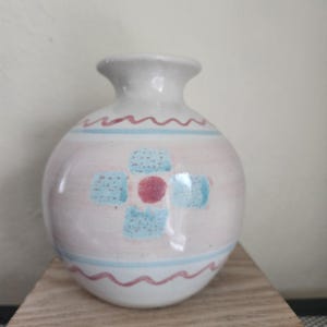May include: A small, round ceramic vase with a narrow neck. The vase is primarily off-white with a pink band and blue and red decorative accents. The design includes a central floral motif and wavy lines.