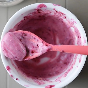 May include: A close-up shot of a white bowl filled with a vibrant pink mixture, possibly a smoothie or yogurt. A pink spoon is partially submerged in the mixture, holding a large scoop. The mixture has a smooth texture with visible dark berry pieces.