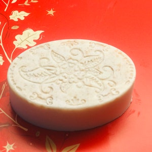 May include: Oval-shaped, beige soap bar with a floral design embossed on the top. The soap is set against a red background with a decorative leaf pattern. The soap appears to be handmade.