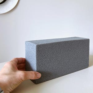 Op de afbeelding: Een rechthoekig blok van grijs schuim met een poreus, getextureerd oppervlak. Het object is solide en heeft een uniforme kleur, met scherpe, strakke randen en een vlakke bovenkant, ontworpen als basis voor knutselwerk of bloemstukken.