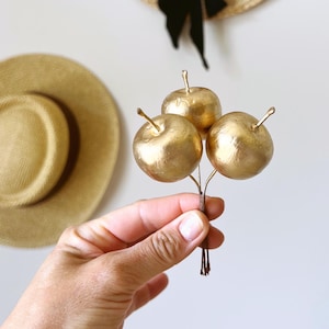 Pode incluir: Um conjunto decorativo de três pequenas maçãs de cor dourada metálica presas a hastes finas de cor castanho-escuro. Cada maçã apresenta uma superfície lisa e arredondada com uma haste curta e pontiaguda que sobressai do topo.