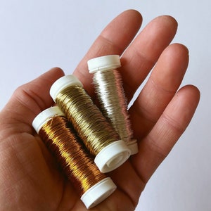 Könnte beinhalten: Drei kleine zylindrische Spulen mit dünnem Metalldraht in Kupfer-, Gold- und Silbertönen. Jede Spule hat weiße Kunststoffkappen an den Enden und ist mit einer durchgehenden Länge aus feinem, reflektierendem Draht für Bastelarbeiten umwickelt.