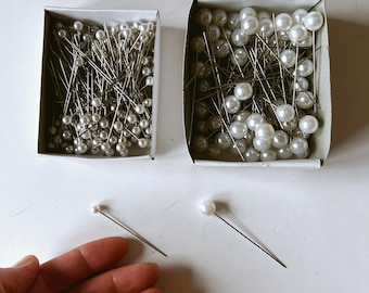 May include: Two cardboard boxes filled with pearl-headed pins. The pins have silver shafts and white, round heads. Two individual pins with pearl heads are also visible. The image is taken from a top-down perspective.