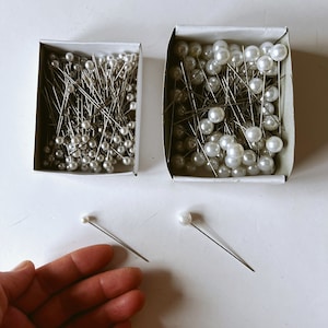 May include: Two cardboard boxes filled with pearl-headed pins. The pins have silver shafts and white, round heads. Two individual pins with pearl heads are also visible. The image is taken from a top-down perspective.