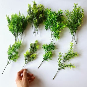 Könnte beinhalten: Sortiment aus zehn künstlichen grünen Pflanzen mit Stielen, angeordnet auf einer weißen Oberfläche. Die Pflanzen sind alle in verschiedenen Formen und Größen, und einige haben ein buschigeres Aussehen als andere.