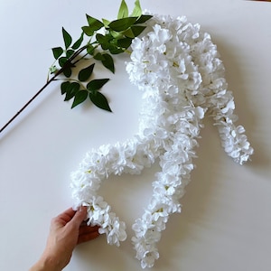 May include: A decorative arrangement of white artificial flowers, similar to wisteria, with a green leafy stem. The flowers are closely packed, giving a full, textured look. The arrangement is set against a white background.