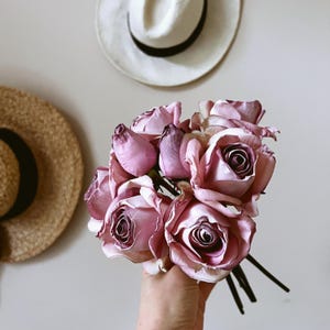 Könnte beinhalten: Ein Strauß malvenfarbener Rosen mit dunklen Zentren und grünen Stielen. Die Rosen werden in einer Hand gehalten, mit zwei Strohhüten im Hintergrund an der Wand.