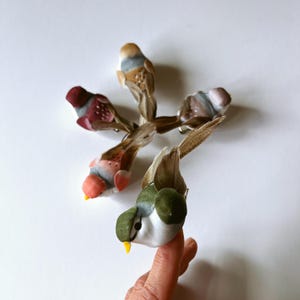 May include: A collection of five small bird ornaments featuring soft, textured bodies in various colors including green, red, brown, and white. Each bird has a pointed yellow beak, dark eyes, and long, thin tail feathers extending outward.