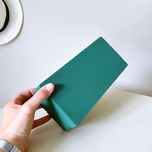 May include: A rectangular block of dark green floral foam with a porous, matte texture. The object is solid and uniform in color, featuring sharp, clean edges and a dense, lightweight appearance typical of materials used for arranging flowers.