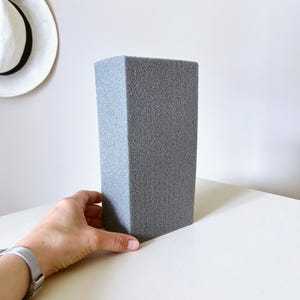 May include: A rectangular block of grey floral foam with a porous, textured surface. The block is solid and uniform in color, featuring sharp, clean edges and a dense, lightweight appearance suitable for various crafting and floral arrangement projects.