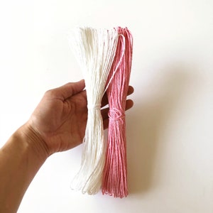 May include: Two bundles of string, one white and one pink, held against a white background. The string is tightly wound and tied in the middle of each bundle. The pink string is a vibrant, light shade.