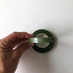 May include: Two rolls of dark green floral tape with a textured, matte finish. The rolls are stacked, with one positioned horizontally and the other vertically, featuring a central white plastic core visible through the center of the tape.
