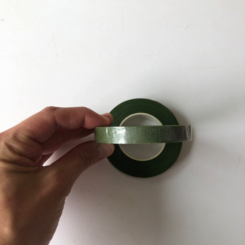 May include: Two rolls of dark green floral tape with a textured, matte finish. The rolls are stacked, with one positioned horizontally and the other vertically, featuring a central white plastic core visible through the center of the tape.