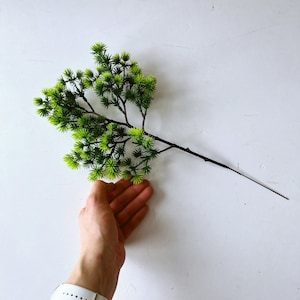 May include: A decorative artificial plant stem featuring a dark brown branch with multiple smaller twigs extending outward. The foliage consists of numerous small, spiky, needle-like leaves in vibrant shades of lime green and deep forest green.