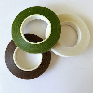 May include: Three rolls of floral tape arranged in a cluster. The rolls are colored in dark green, dark brown, and off-white. Each roll features a textured surface and a hollow center, showing the white plastic core used for winding the tape.
