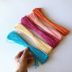 May include: Bundles of colorful twine in shades of turquoise, cream, magenta, peach, and orange are arranged on a white surface. The twine is tightly wound and tied, ready for crafting or decorative use. The image is well-lit, showcasing the vibrant colors.