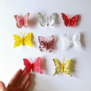 May include: A collection of decorative butterfly clips in various colors, including red, yellow, white, and pink. The butterflies have different patterns and textures, some with glitter accents. They are arranged on a white surface.