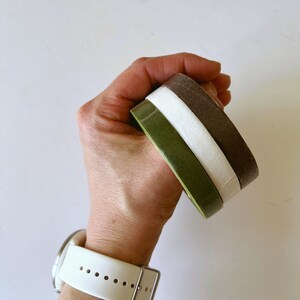 May include: Three stacked circular bands in olive green, white, and dark brown. The bands have a smooth, solid finish and are held together in a stack. A white silicone watch band with a silver buckle is visible on the wrist.